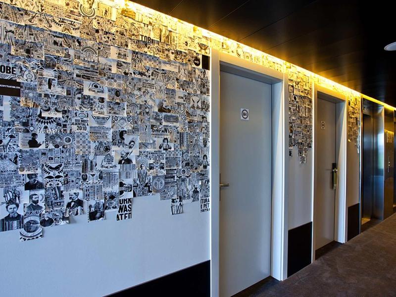 Flur mit weißen Türen, beleuchtet durch gelbes LED-Licht, und Wand mit schwarz-weißen Fotos und Zeichnungen.