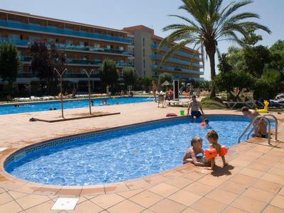 Bambini che giocano in una piscina bassa davanti a un hotel con giardino e palme.