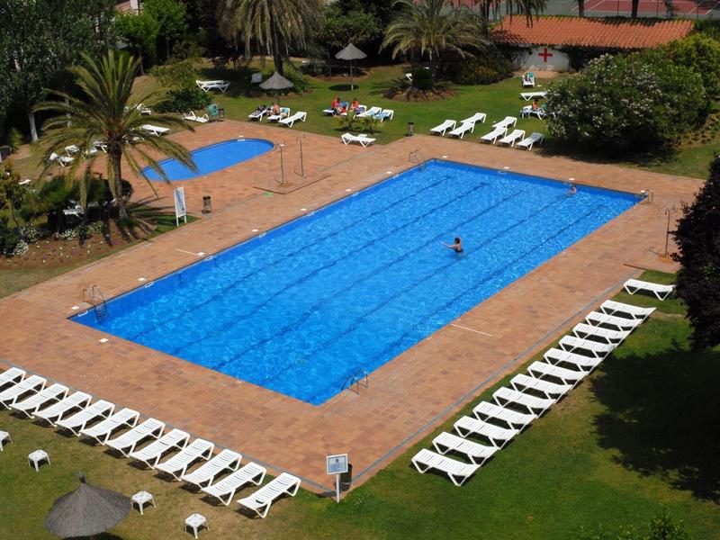 Una grande piscina rettangolare con acqua blu, circondata da sedie a sdraio bianche in un giardino d'hotel.