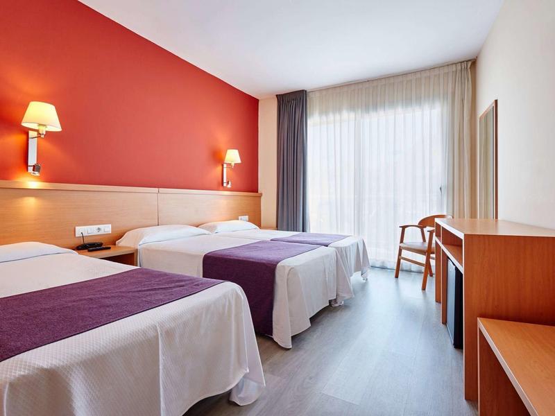 Camera d'hotel con due letti singoli, parete rossa, scrivania e finestra con tende.