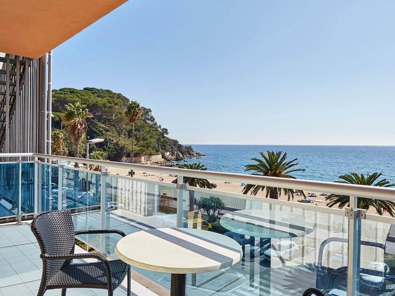 Balcone con due sedie e tavolo con vista sul mare e palme sotto un cielo sereno.