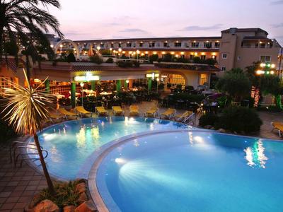 Hotel mit beleuchtetem Pool, Palmen und Sitzbereichen bei Dämmerung, warme Atmosphäre.