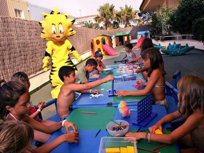 Kinder sitzen an einem langen Tisch im Freien und spielen Brettspiele, begleitet von zwei verkleideten Figuren.