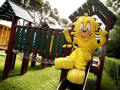 Gelbe Plüschfigur sitzt fröhlich auf Spielplatz-Rutsche aus Holz und Metall im Grünen.
