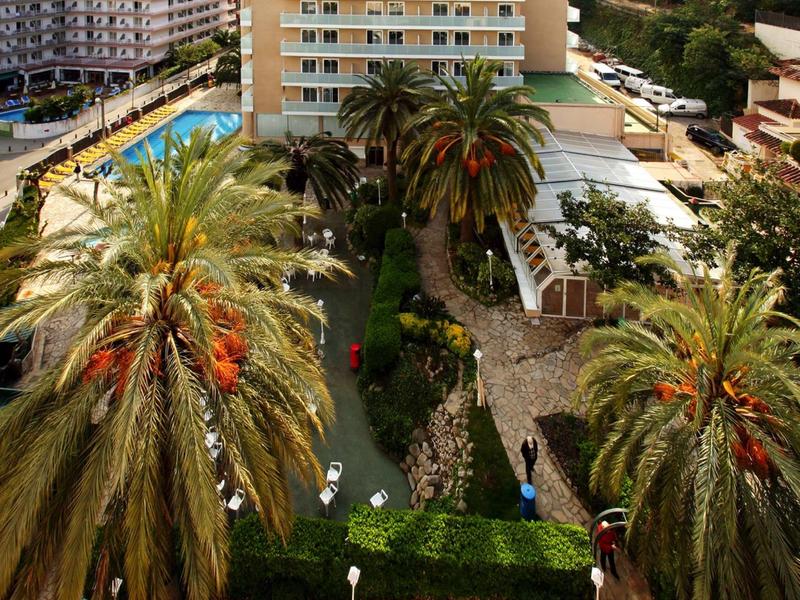 Budova hotelu s bazénem, palmami a oplocenou zahradou v městském prostředí.