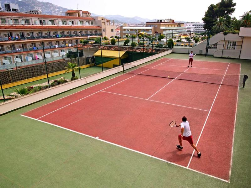 Dva hráči na červeném tenisovém dvorci vedle hotelové budovy za slunečného počasí.