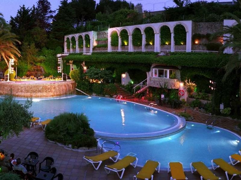 Noční venkovní hotelový bazén s osvětlenými fontánami a lehátky.