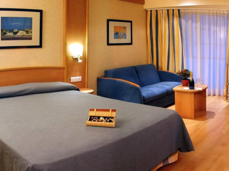 Pohodlný hotelový pokoj s velkou postelí, pohovkou a balkonem, teplé tóny.