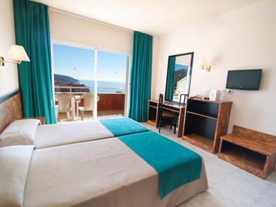 Hotelzimmer mit zwei Betten, türkisfarbenen Vorhängen, Schreibtisch, Fernseher und Balkon mit Meerblick.