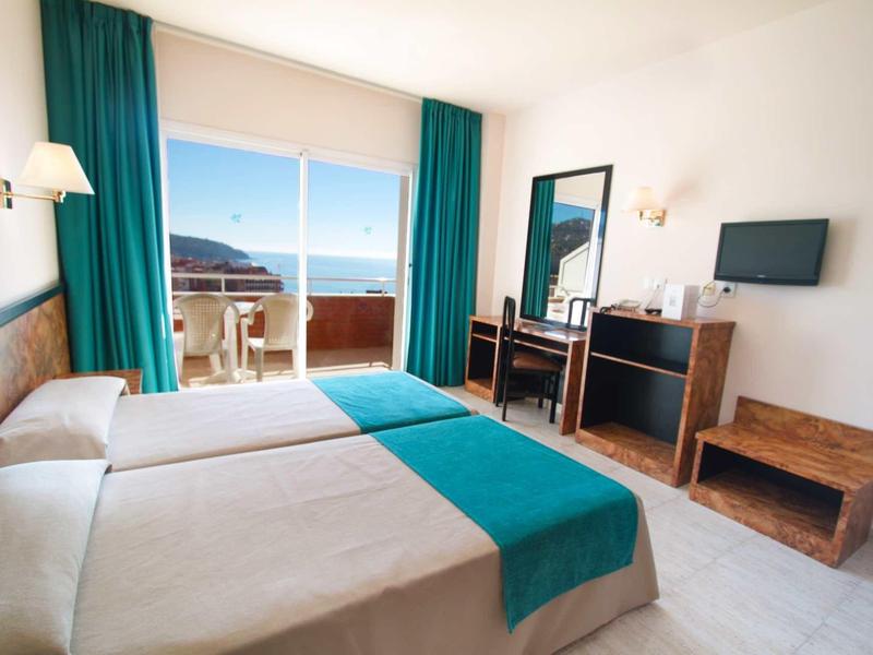 Hotelzimmer mit zwei Betten, türkisfarbenen Vorhängen, Schreibtisch, Fernseher und Balkon mit Meerblick.