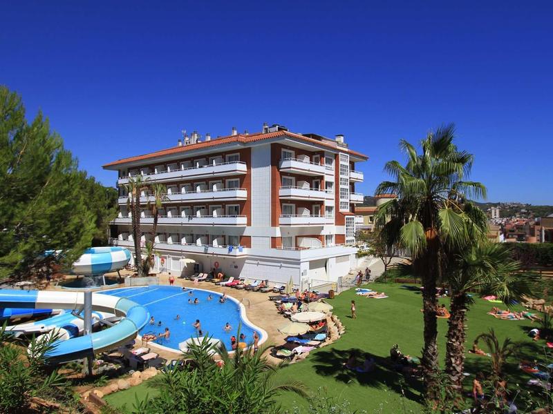 Mehrstöckiges Hotelgebäude mit Pool, Sonnenschirmen, Palmen und grünem Garten unter blauem Himmel.