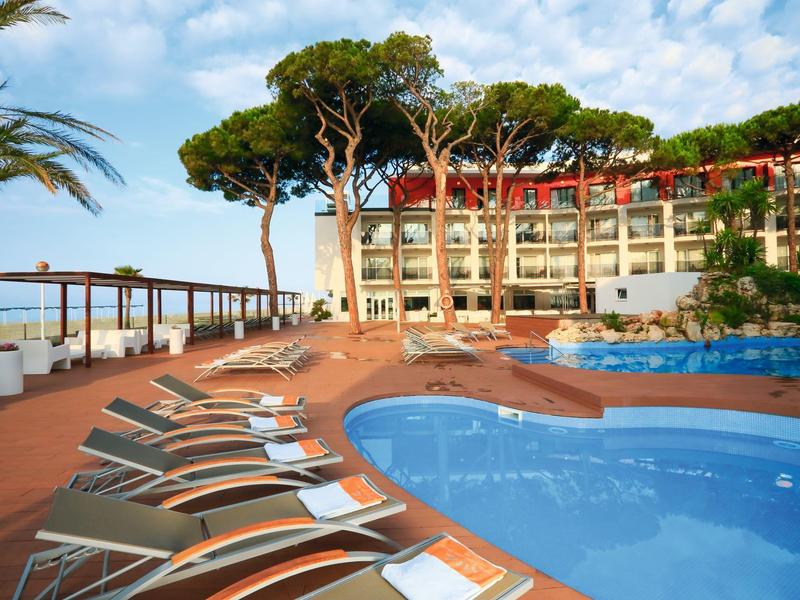 Resort pool area with lounge chairs, umbrellas, trees, and a hotel building in the background.