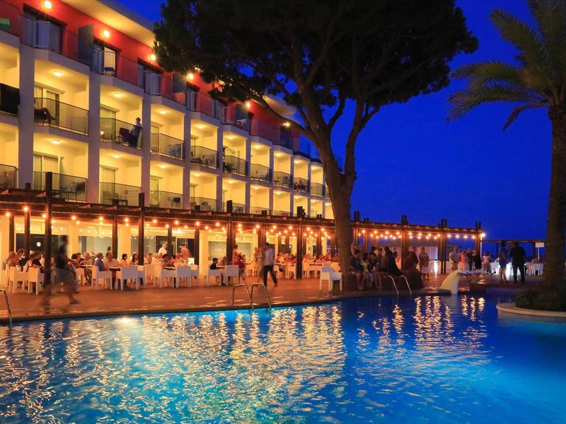 Hotel pool and terrace lit at night with palm trees and people dining outdoors.