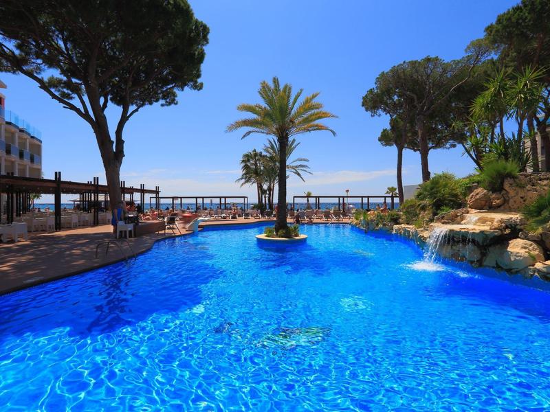 Bright blue pool with palm tree centerpiece, surrounded by trees and seating areas near the sea.