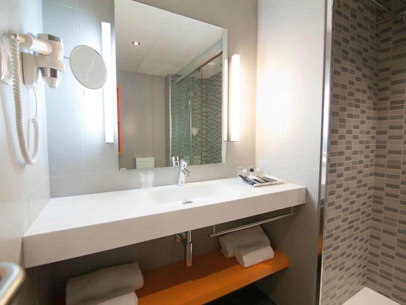 Modern hotel bathroom with large mirror, white countertop, sink, hairdryer, and tiled shower area.