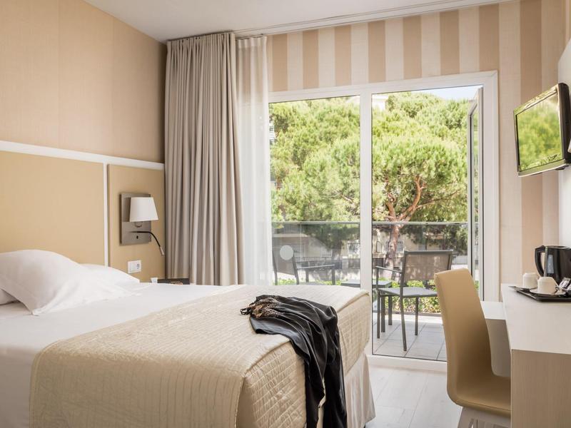 Bright hotel room with double bed, desk, chair, TV, and balcony with trees outside.