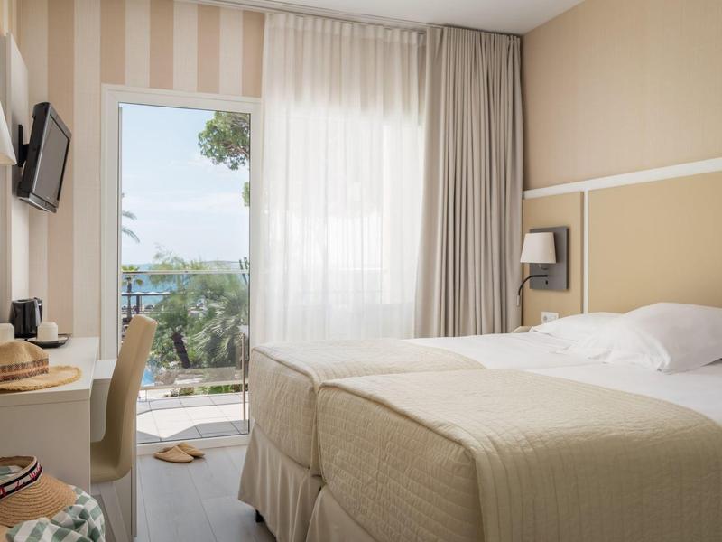 Bright hotel room with double bed, wall-mounted TV, desk, and balcony with outdoor view.