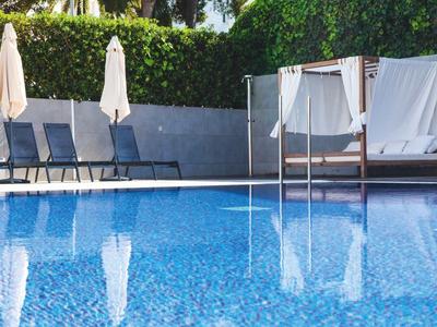 Ein ruhiger Pool mit blauer Wasseroberfläche, Liegestühlen, weißen Sonnenschirmen und grüner Hecke.