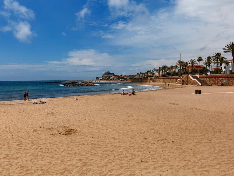 Weicher Sandstrand mit blauem Himmel, Palmen und wenigen Menschen am Wasser.