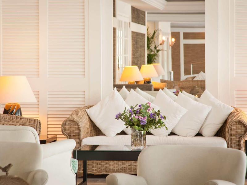 Salon d'hôtel confortable avec fauteuils en osier, coussins blancs et bouquet sur la table.