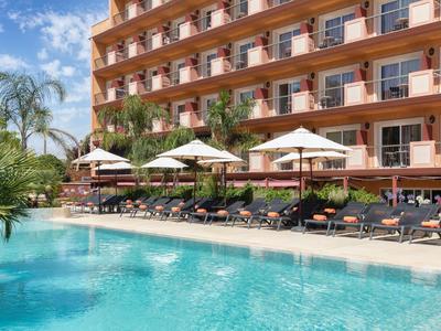 Hotelgebäude mit Balkonen, Palmen und Pool mit Liegestühlen unter Sonnenschirmen im Außenbereich.