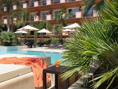 Piscina del hotel con tumbonas, sombrillas y palmeras, rodeada de un edificio de varias plantas.