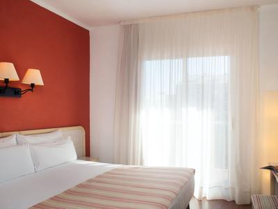 Habitación de hotel luminosa con cama doble, pared roja y ventana grande con cortinas.