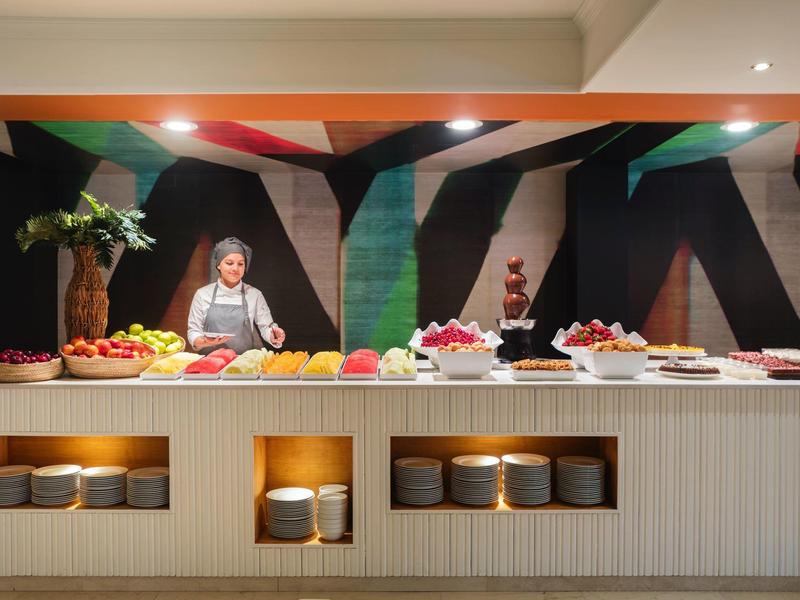 Buffet con frutas y postres en un restaurante de hotel moderno, camarera al fondo.