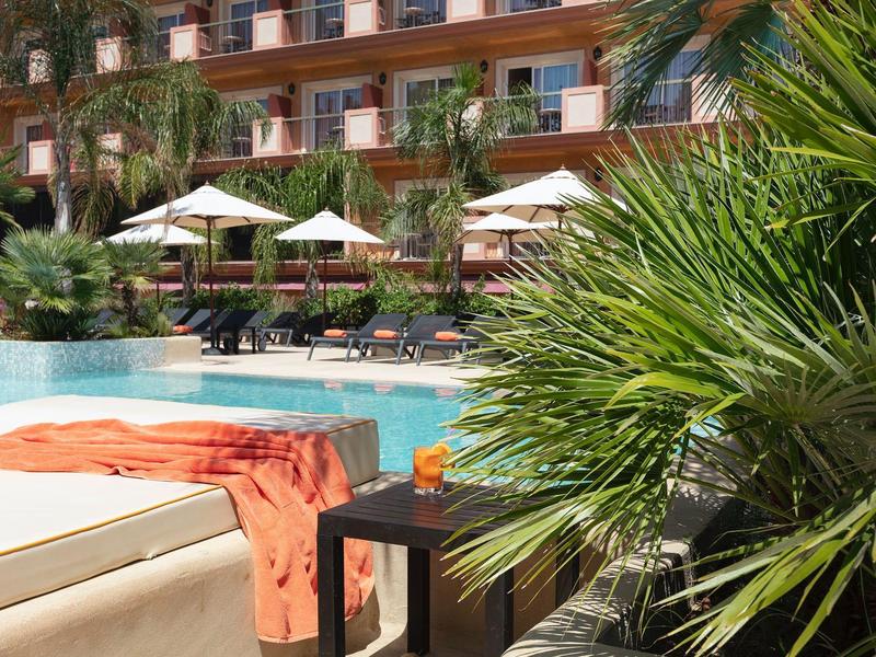 Piscina del hotel con tumbonas, sombrillas y palmeras, rodeada de un edificio de varias plantas.