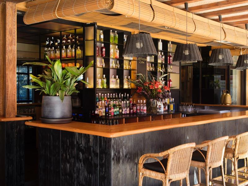 Área moderna de bar de hotel con barra de madera, taburetes y estantes iluminados de bebidas.