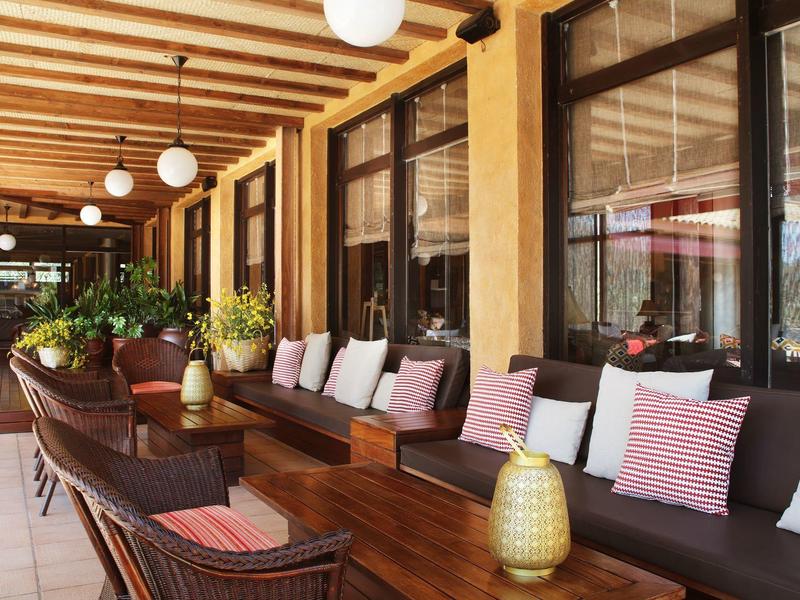 Cómodo espacio exterior con muebles de madera, cojines y plantas verdes bajo vigas de madera.