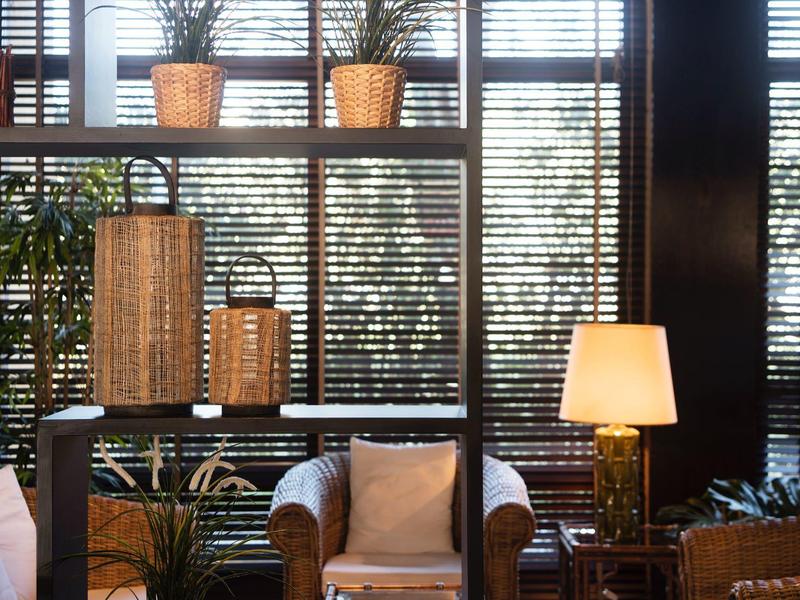 Acogedora sala de hotel con plantas, lámparas y grandes ventanas con persianas a la luz del día.