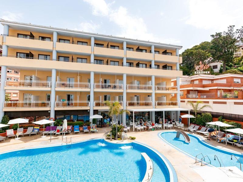 Complejo hotelero moderno de varias plantas con terrazas y gran piscina al aire libre bajo cielo azul.