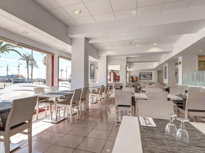 Comedor moderno y luminoso con filas de mesas y sillas y vista a palmeras y el mar.