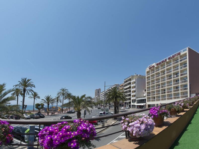 Hoteles modernos con palmeras y flores coloridas bajo un cielo azul claro en zona turística.