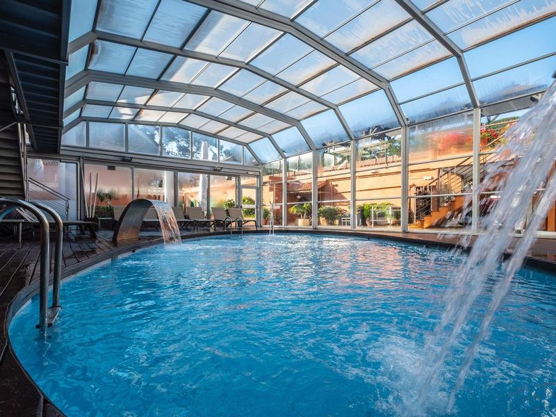 Piscina cubierta con techo acristalado y cascada de agua en un entorno de hotel.