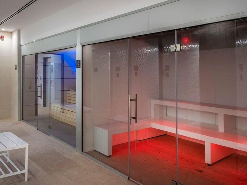 Área de sauna moderna con paredes de cristal, bancos blancos y iluminación roja en el suelo.