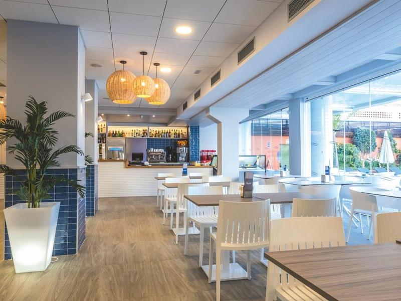 Área luminosa del restaurante del hotel con suelo de madera, muebles blancos y grandes ventanales a la terraza.