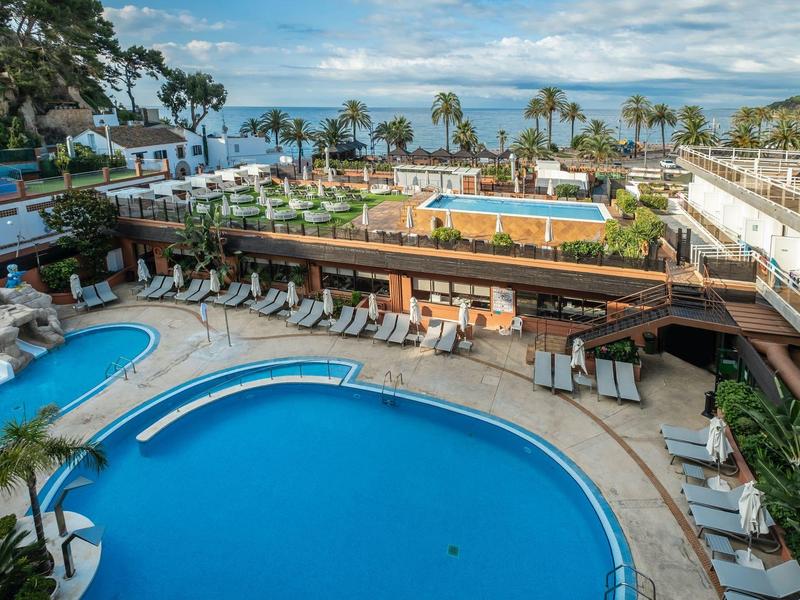 Gran piscina de hotel con tumbonas, zona de bar y vista al mar con palmeras al fondo.