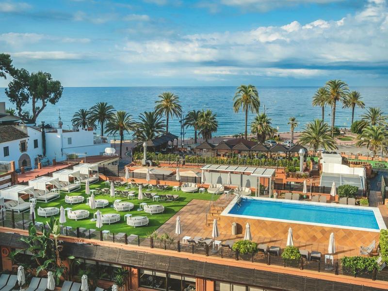 Vista de un jardín de hotel con piscina, palmeras y mar al fondo bajo cielo nublado.
