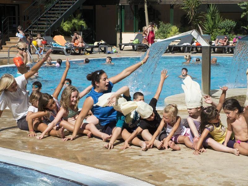 Niños y adultos juegan con pelotas blancas junto a una piscina bajo el sol