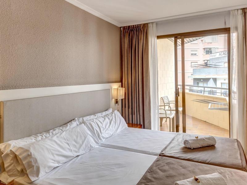 Habitación con dos camas, lámpara de noche y acceso a un balcón con vista a edificios.