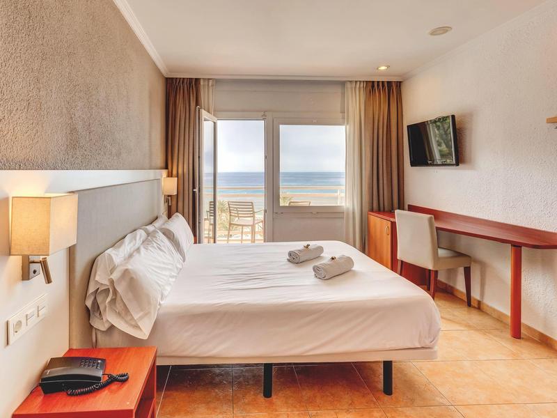 Habitación de hotel luminosa con cama doble, vista al mar, escritorio y televisor en la pared.