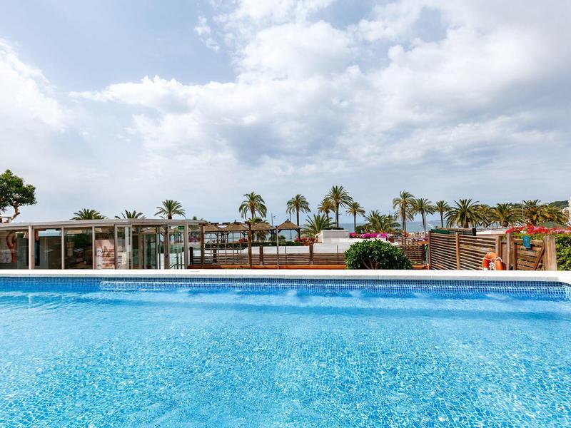 Gran piscina con agua cristalina frente a una fila de palmeras y edificios bajo un cielo nublado.