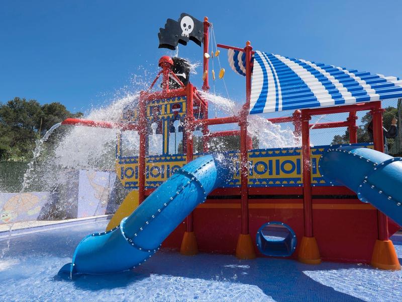 Kolorowy plac zabaw wodny z zjeżdżalniami i motywem statku pod niebieskim niebem.