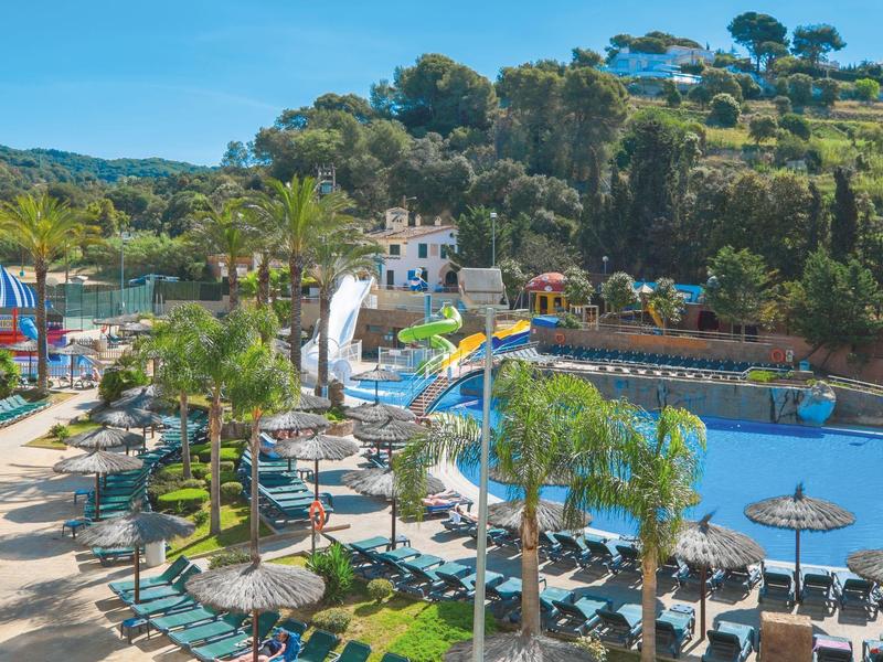 Widok na basen hotelowy z zjeżdżalniami, leżakami i zielonymi wzgórzami w tle.