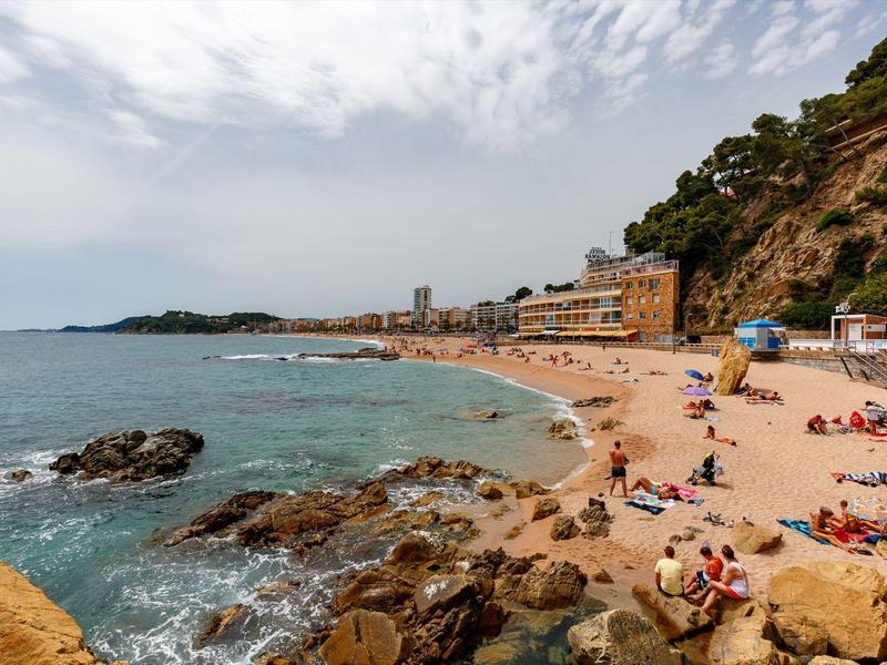 Plaża z skałami, ludźmi i budynkami u podnóża wzgórza pod pochmurnym niebem.