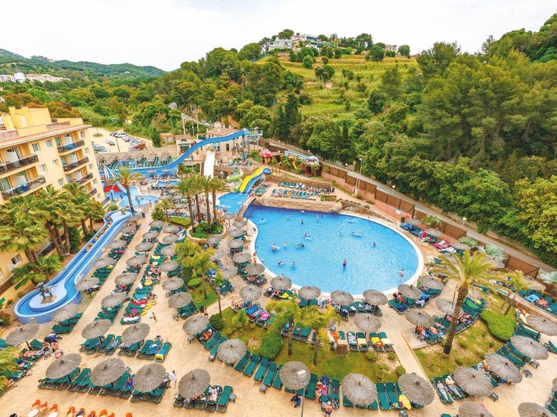 Duży basen hotelowy ze zjeżdżalnią, leżakami i zalesionymi wzgórzami w tle.