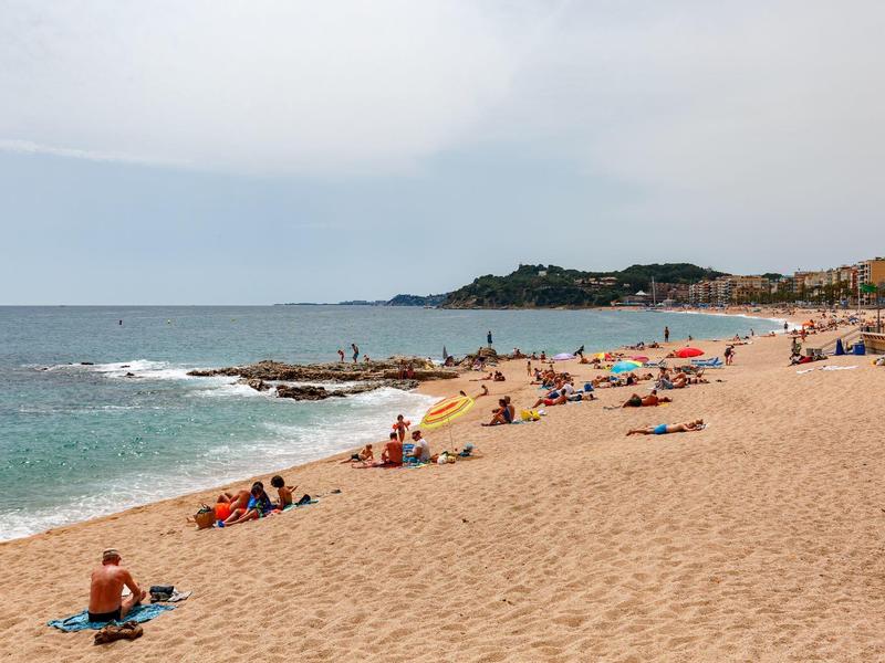 Szeroka piaszczysta plaża z kąpiącymi się i spokojnym morzem pod zachmurzonym niebem.