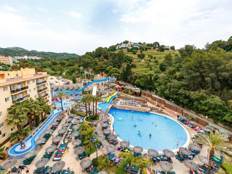 Hotel z dużym basenem zewnętrznym, zjeżdżalnią wodną i zielonym pagórkowatym krajobrazem.